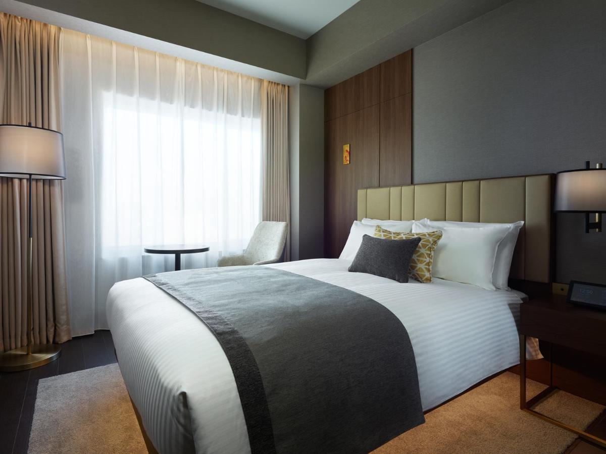 Mitsui Garden Hotel Roppongi Tokyo Premier Экстерьер фото The photo shows a modern hotel room featuring a neatly made bed with white linens and a decorative gray throw blanket. To the side, there is a small round table and a comfortable chair, creating a cozy seating area. The room is well-lit with natural 