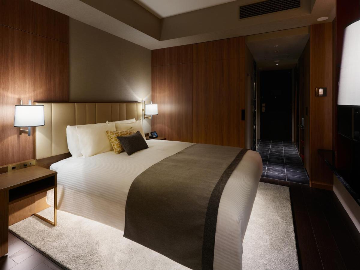 Mitsui Garden Hotel Roppongi Tokyo Premier Экстерьер фото The image shows a modern hotel room with a warm and elegant design. In the center, there is a neatly made bed with white linens and a dark gray throw blanket. Flanking the bed are two bedside lamps providing soft lighting. The walls have a rich wood 