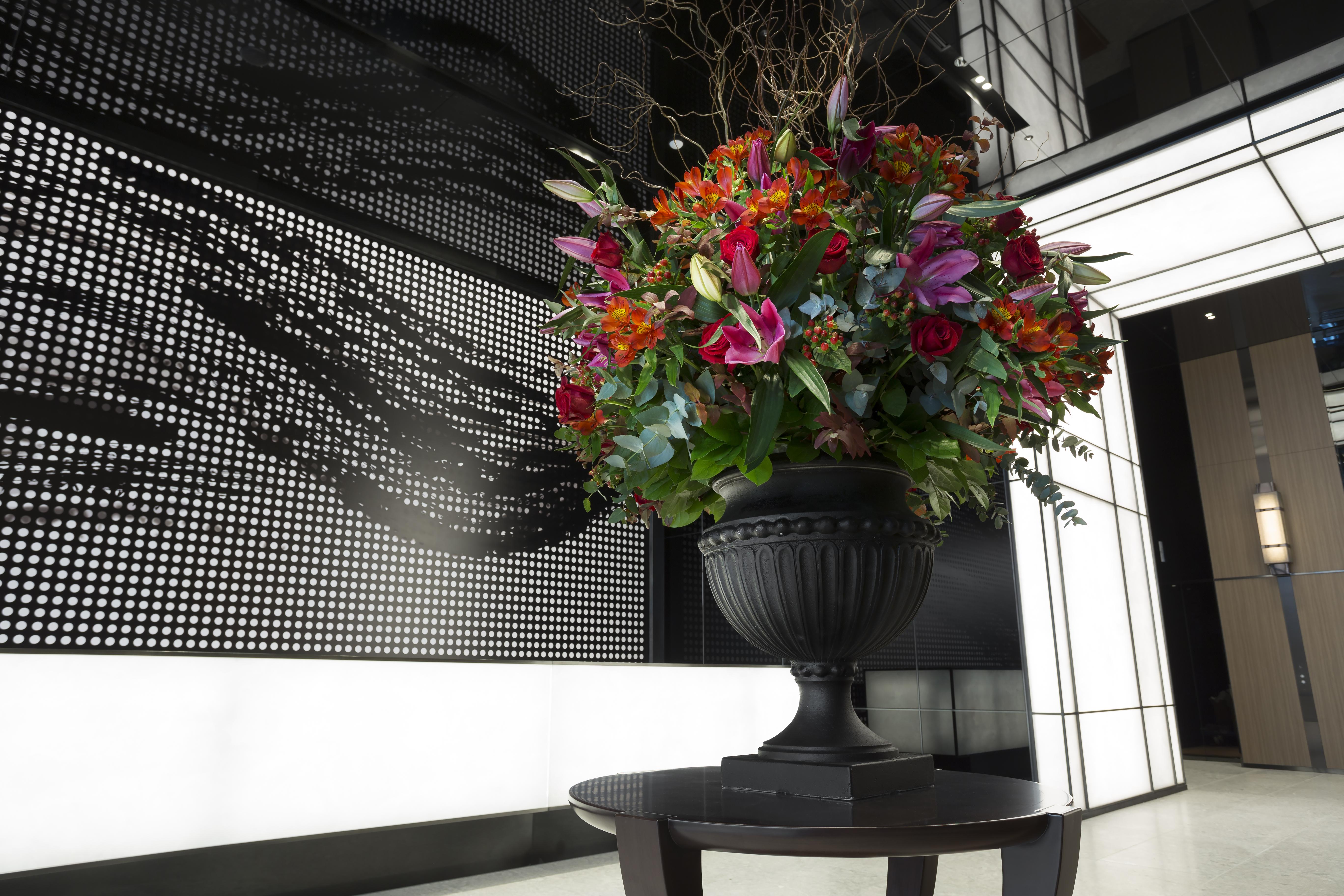 Mitsui Garden Hotel Roppongi Tokyo Premier Экстерьер фото The photo features a large, vibrant floral arrangement in an ornate black urn. The arrangement is composed of various colorful flowers and greenery, showcasing shades of red, pink, orange, and green. This floral display is positioned on a small table
