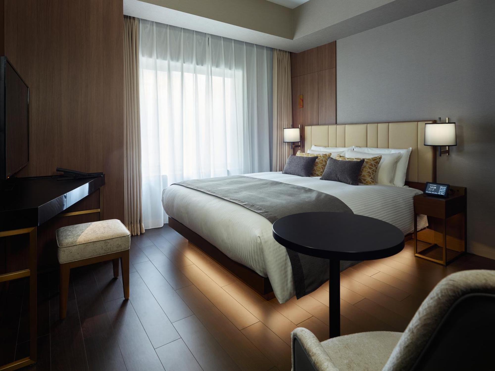 Mitsui Garden Hotel Roppongi Tokyo Premier Экстерьер фото The photo shows a modern hotel room featuring a large bed with decorative pillows. There are large windows with sheer curtains allowing natural light to filter in. To one side, there is a sleek desk with a chair and a small round table in the center 