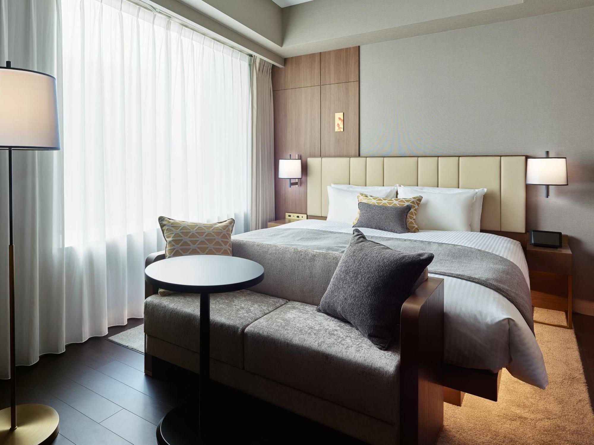 Mitsui Garden Hotel Roppongi Tokyo Premier Экстерьер фото The photo shows a modern hotel room that features a large bed with a padded headboard and soft bedding. There's a small, round table accompanied by a couch or seating area, adorned with decorative pillows. To the left, a floor lamp provides additiona